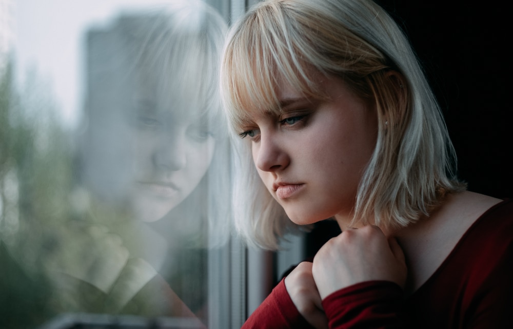 Depressed young woman near window illustrating the challenges of addiction substance abuse and the need for rehabilitation