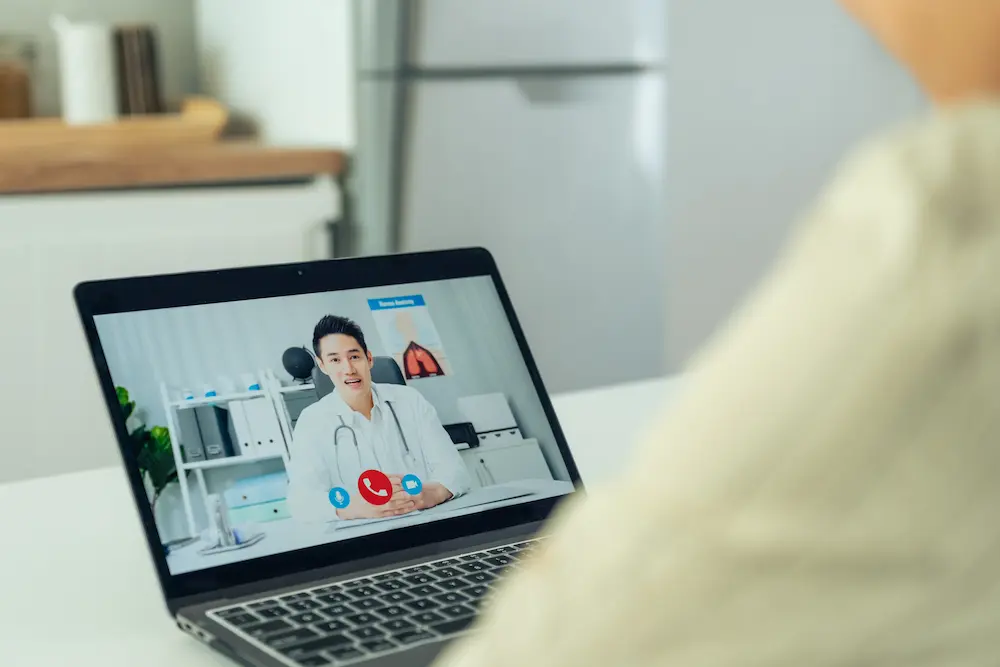An over-the-shoulder view of a person looking at a laptop screen displaying a video consultation with a doctor. This illustrates telehealth services, which are mentioned as an accessible option for obtaining medication-assisted treatment, such as buprenorphine, for tianeptine use disorder.