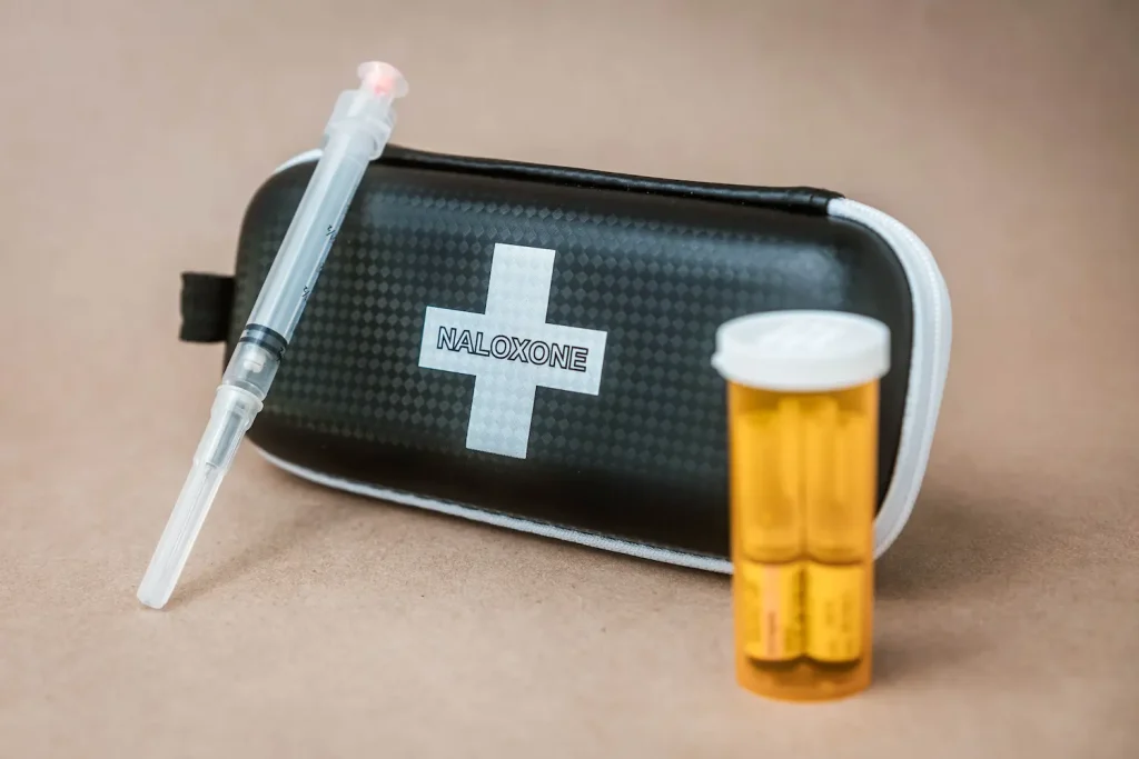 A black zippered carrying case with a white cross and the word 'NALOXONE' printed in the center. To the left of the case rests a clear medical syringe, and to the right is an amber prescription bottle containing two small glass vials. The items are displayed on a plain light brown surface.