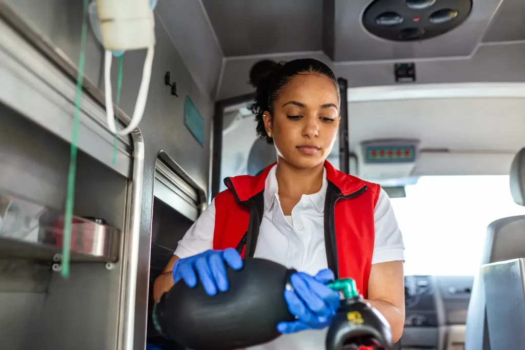 A female emergency medical technician (EMT) standing inside an ambulance. She is wearing a white polo shirt, a red vest, and blue medical gloves, and is holding and preparing a black bag valve mask (BVM) used for manual resuscitation and breathing support.