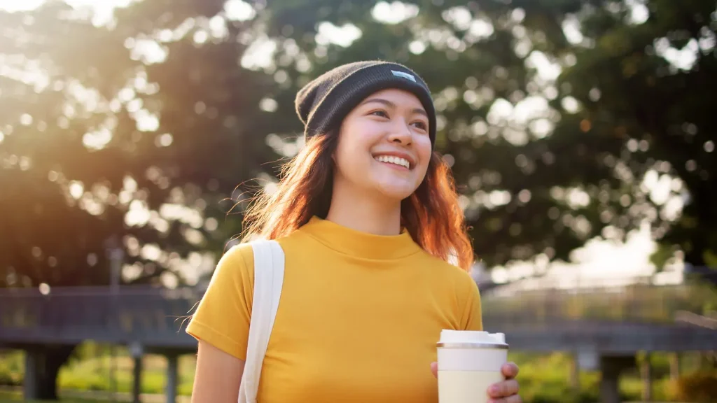 A smiling young woman wearing a yellow shirt and beanie, walking outdoors with a coffee cup.