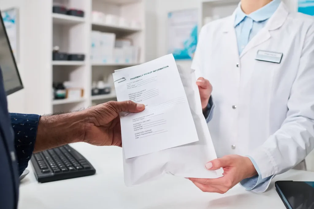 A customer receiving a prescription bag from a pharmacist over a white counter.