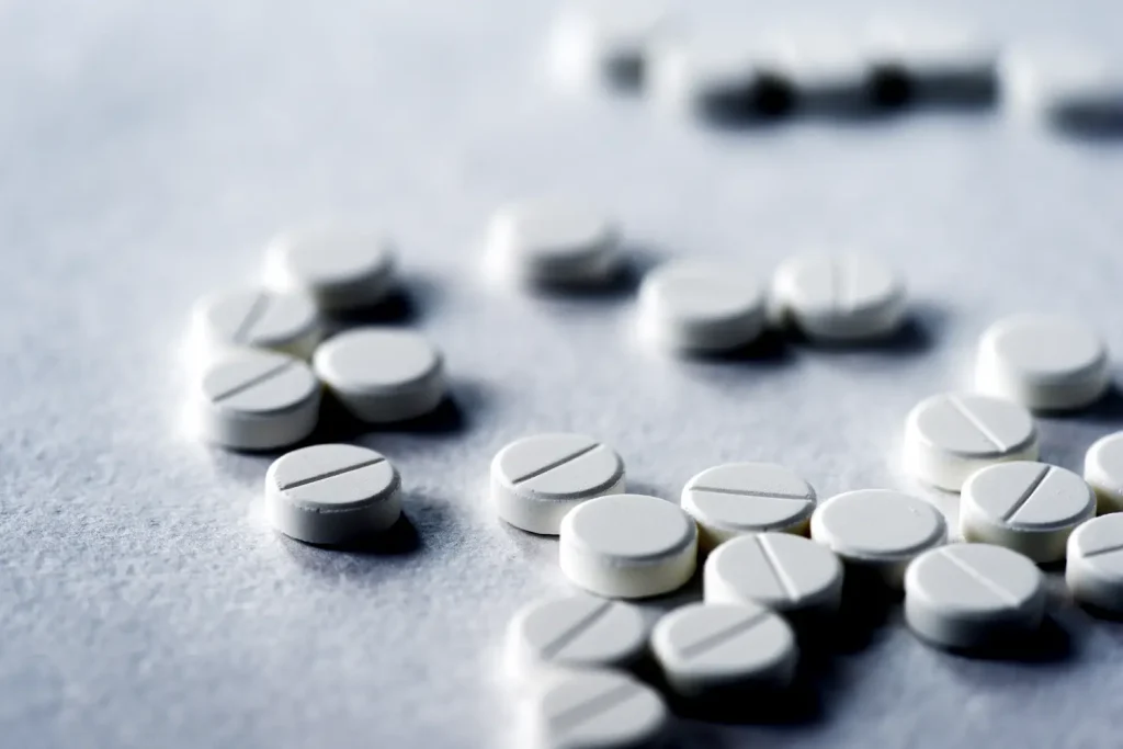 A close-up of several round, white pills with a single score line down the middle, scattered across a light, textured surface.