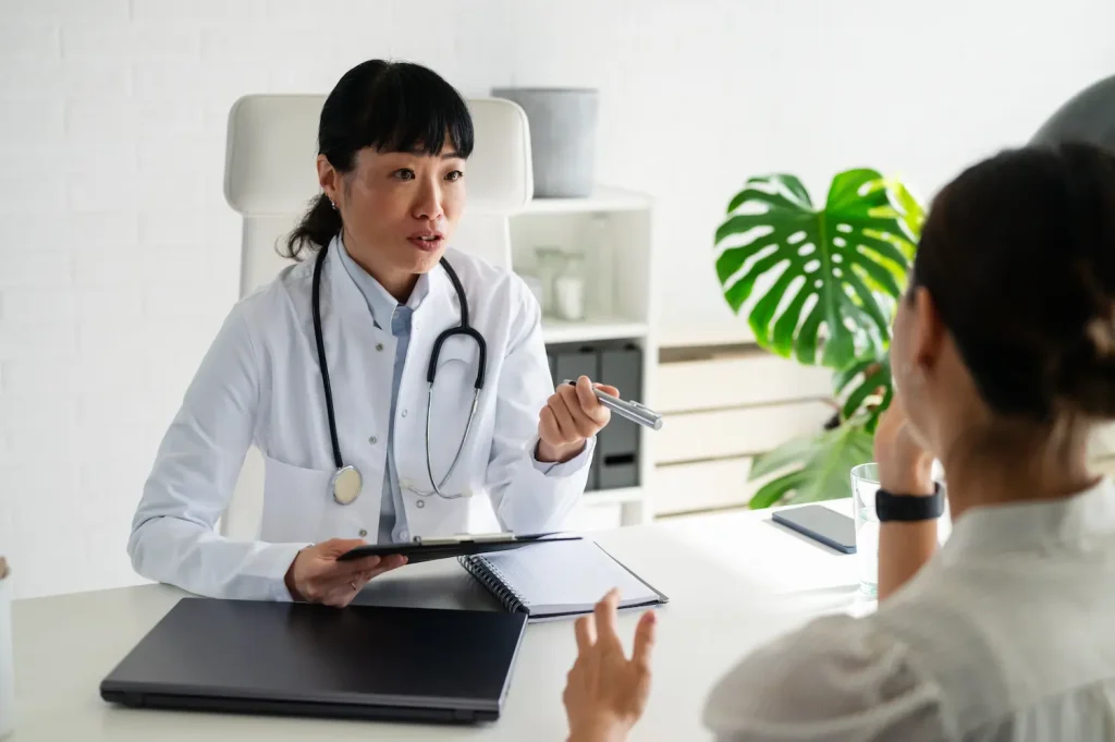 A female doctor wearing a white lab coat and stethoscope sits at a desk, holding a pen and clipboard as she speaks with a patient seated across from her in a brightly lit clinic office.