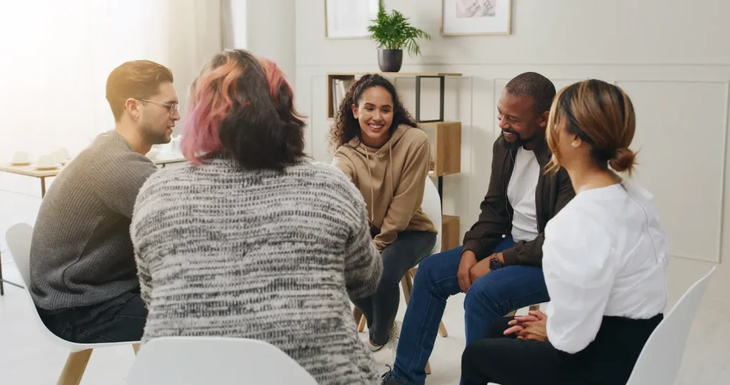 a diverse group of five adults sits in a circle on white chairs in a bright, modern room, resembling a support group or group therapy session. In the center, a smiling woman with curly hair wearing a beige hoodie is looking toward the others, appearing engaged in a positive conversation. The other members—including a man with glasses on the left and a smiling man in a dark jacket on the right—listen attentively. The backs of two individuals (one with pink-streaked hair and a grey sweater, the other in a white shirt) are visible in the foreground, framing the group dynamic.