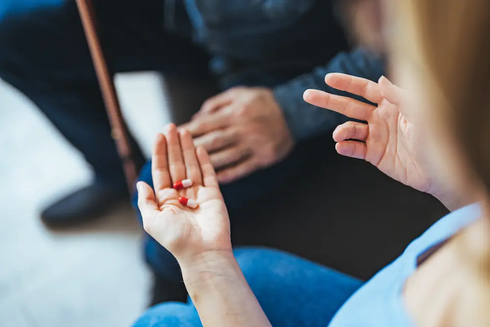 Close-up of a woman holding two red-and-white pill capsules in her open palm while gesturing with her other hand.