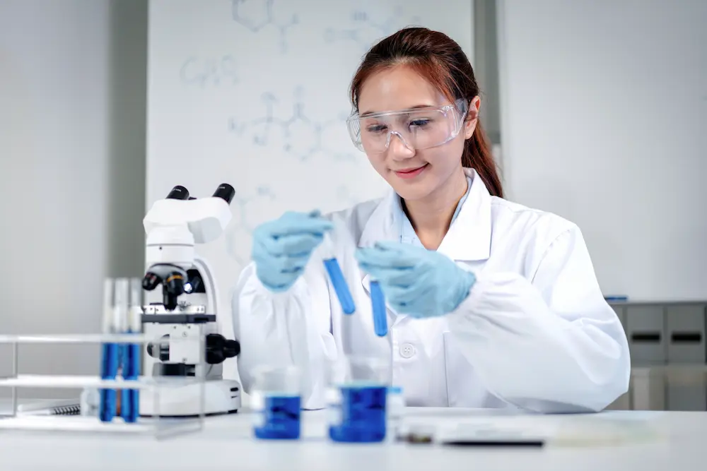 A female scientist in safety goggles and blue gloves handling test tubes filled with blue liquid in a laboratory setting.